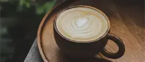 Ein Holzteller hält eine Kaffeetasse mit Latte-Art. Das Latte zeigt ein Herzdesign auf der Oberseite. Die Umgebung ist im Freien mit sichtbarer Grünfläche im Hintergrund.