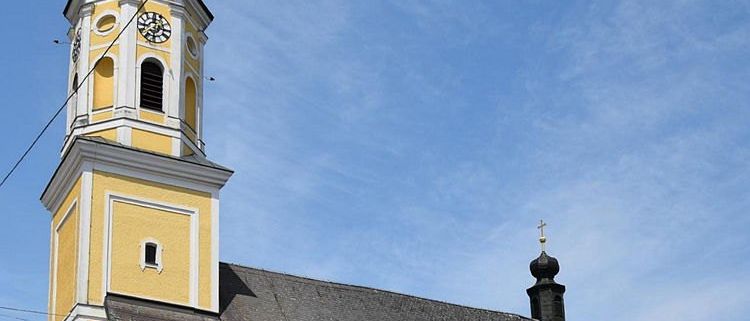 Eine gelbe Kirche mit einem grauen Dach und einem Turm mit einer Uhr. Die Kirche befindet sich in einer Stadt mit blauem Himmel.