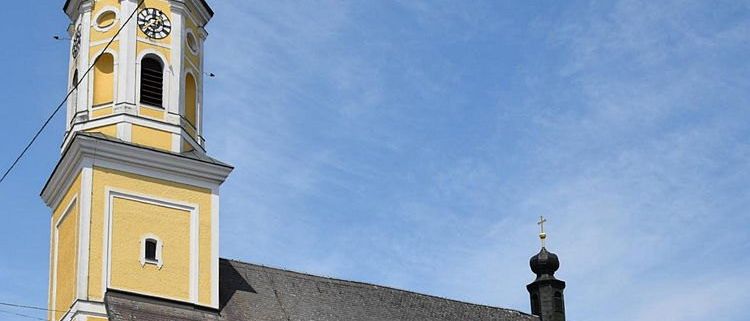 Eine gelbe Kirche mit einem grauen Dach und einem hohen Turm. Der Turm hat eine Uhr und ein Kreuz auf der Spitze. Die Kirche hat mehrere bogenförmige Fenster und eine Statue davor.