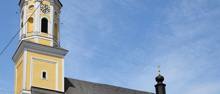 Eine gelbe Kirche mit einem grauen Dach und zwei Türmen. Ein Turm hat eine Uhr, der andere einen Kreuz. Es gibt mehrere Bogenfenster am Gebäude.