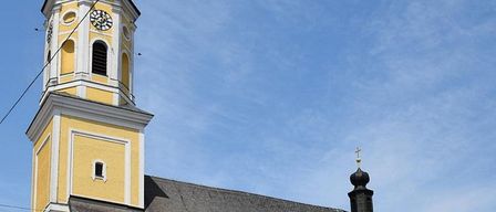 Eine gelbe und weiße Kirche mit einem Turm, einer Uhr und einem Kreuz. Das Gebäude hat ein graues Dach und mehrere bogenförmige Fenster. Es befindet sich unter einem klaren blauen Himmel.