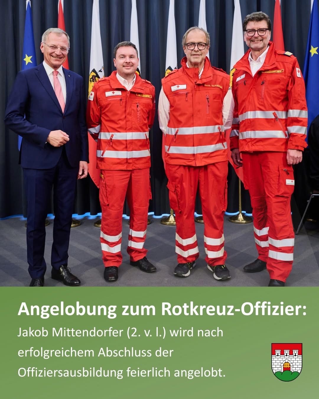 Vier Männer in roten Uniformen stehen zusammen, einer in einem Anzug. Hinter ihnen sind Fahnen. Der Text lautet: 'Ernennung zum Rotkreuz-Offizier: Jakob Mittendorfer (2. v. l.) wird nach erfolgreichem Abschluss der Ausbildung ernannt.'