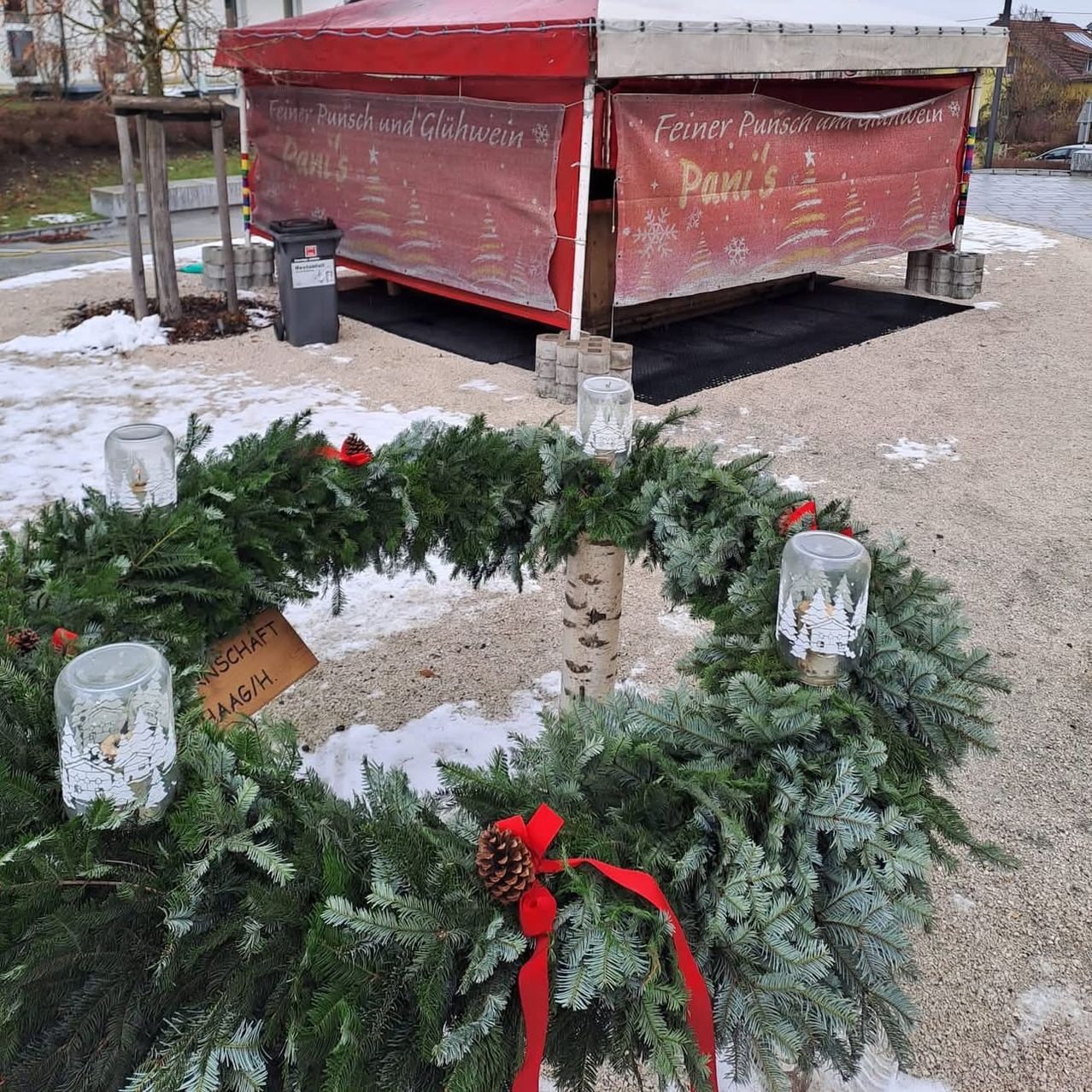Ein Weihnachtskranz schmückt einen Baum vor einem roten Stand, der Glühwein verkauft. Schnee bedeckt den Boden und ein Mülleimer ist in der Nähe.