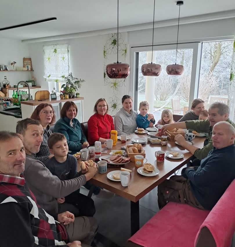 Eine große Gruppe von Menschen sitzt um einen Esstisch herum und genießt eine Mahlzeit. Sie lächeln und scheinen in fröhlicher Stimmung zu sein.