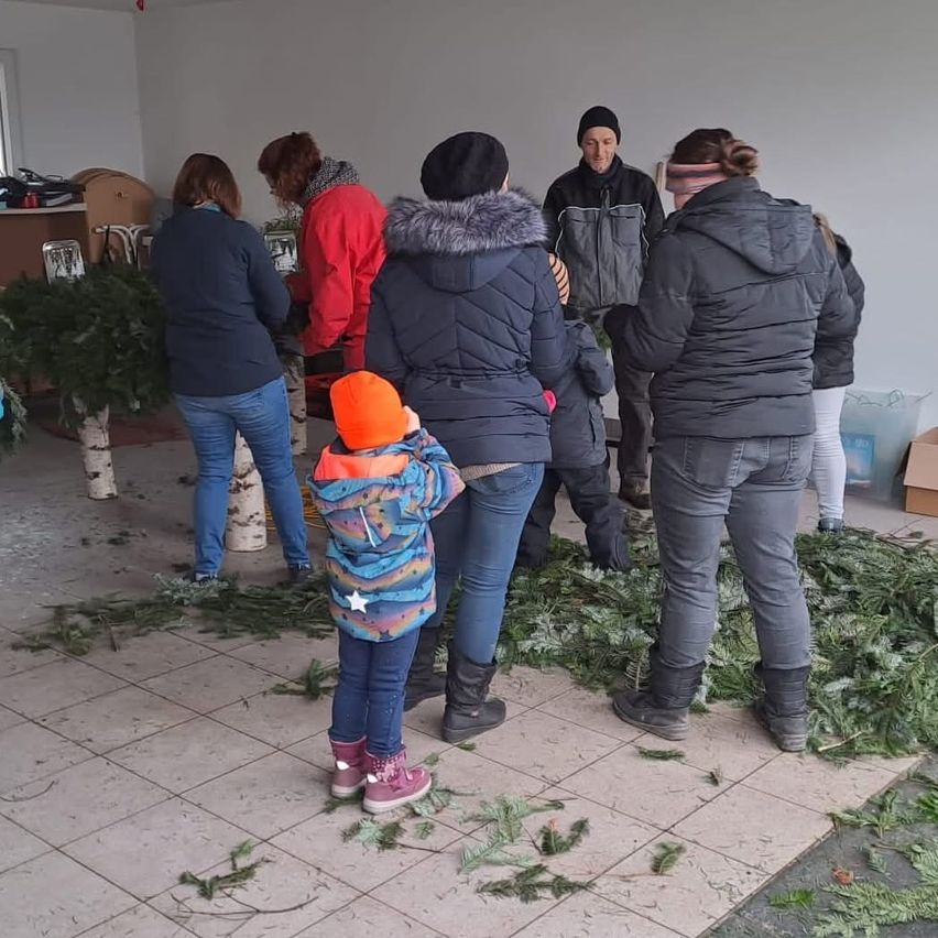 Eine Gruppe von Menschen ist drinnen, wahrscheinlich in einer Fabrik, umgeben von Tannenzweigen. Ein Kind in einem farbenfrohen Mantel steht vor einem Haufen Zweige.