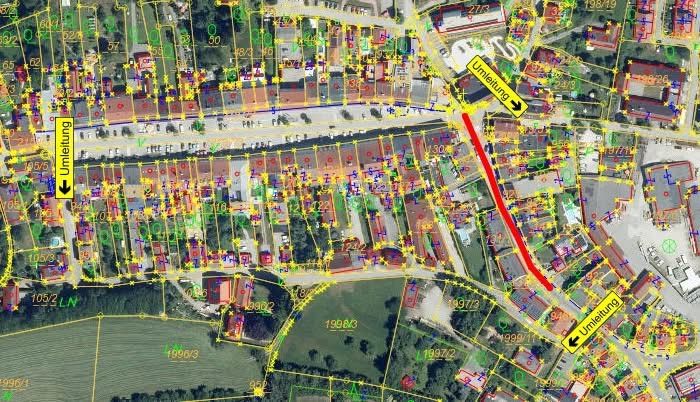 Die Luftaufnahme zeigt Wohngebäude mit gelben Linien, einschließlich einer roten Linie, die eine Hauptstraße anzeigt. Die Karte enthält gelbe Pfeile, die auf Orte zeigen, die als 'Umleitung' gekennzeichnet sind. Zahlen und Grundstücke sind mit verschiedenen Bezeichnungen und Farben gekennzeichnet.