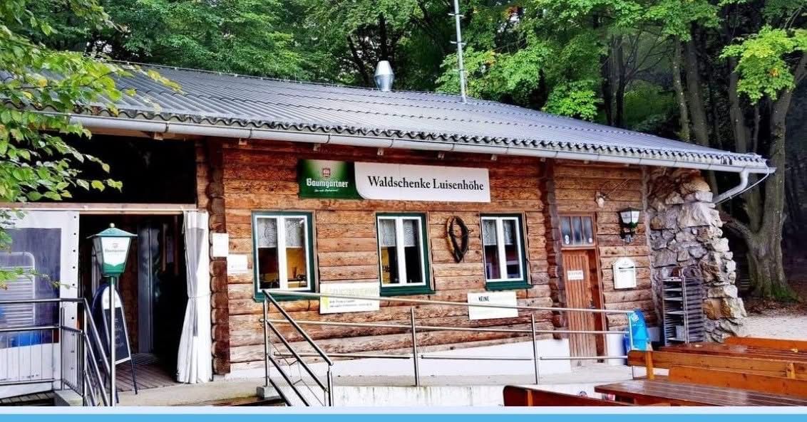 Ein Holzhaus mit Metalldach. Es hat ein Schild mit der Aufschrift Waldschenke Luisenhöhe und drei Fenster. Ein Metallgeländer führt zum Eingang. Bäume stehen hinter dem Gebäude.