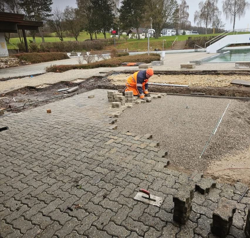 Ein Bauarbeiter in oranger Schutzhelm und Sicherheitsweste arbeitet auf einem gepflasterten Gebiet. Es gibt Kies und Pflastersteine auf dem Boden.