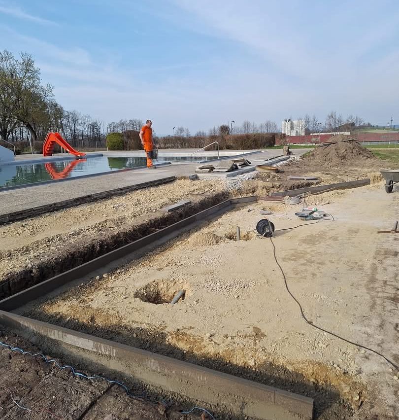 Eine Baustelle in der Nähe eines Pools mit einem Arbeiter in oranger Arbeitskleidung. Der Pool hat eine rote Rutsche und der Boden ist mit Erde gefüllt. Bäume und Gebäude sind im Hintergrund.