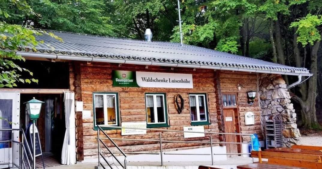 Eine Blockhütte mit grauem Metalldach und Holzwänden. Das Schild steht Waldschenke Luisenhoehe. Die Fenster haben weiße Vorhänge. Eine Brüstung und eine Tür befinden sich auf der rechten Seite. Bäume sind im Hintergrund.