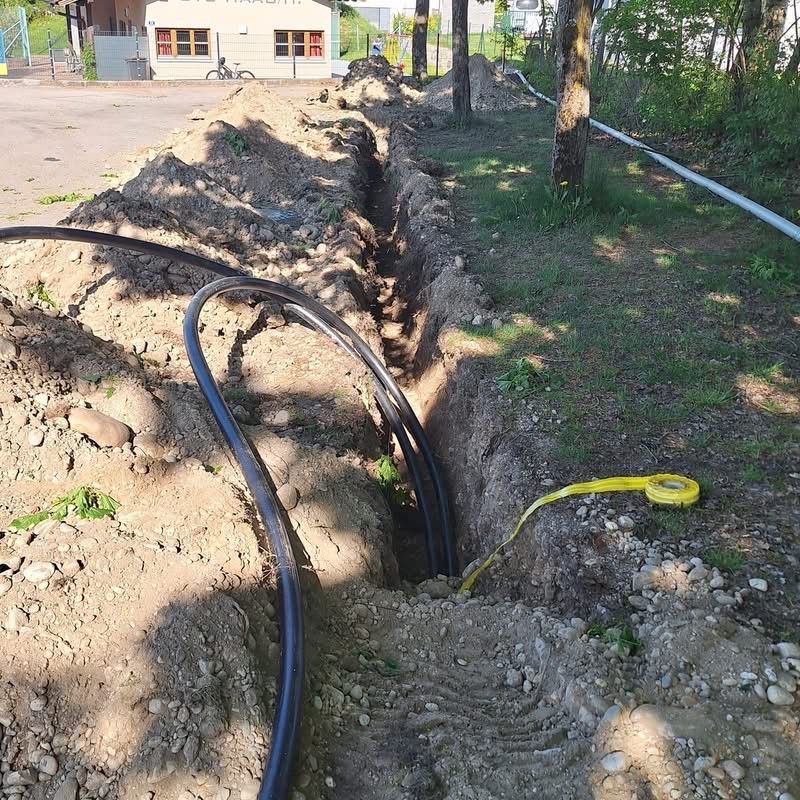 Eine offene Grube in einem Park mit zwei schwarzen Rohren und einem gelben Messband auf dem Boden. Ein Haus mit einem Fahrrad davor und Bäume im Hintergrund.