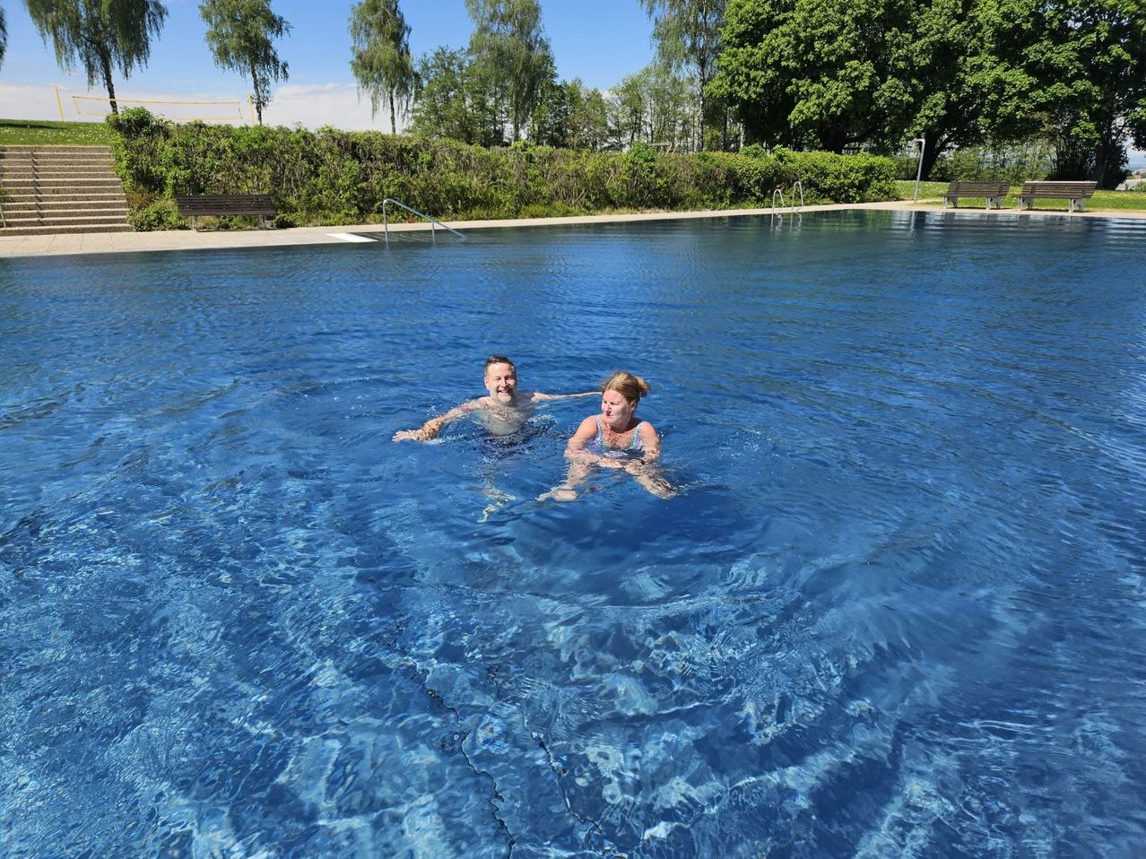 Zwei Personen, ein Mann und eine Frau, schwimmen in einem großen Freibad mit klarem blauem Wasser und einer Betonterrasse. Bäume und Büsche sind im Hintergrund.