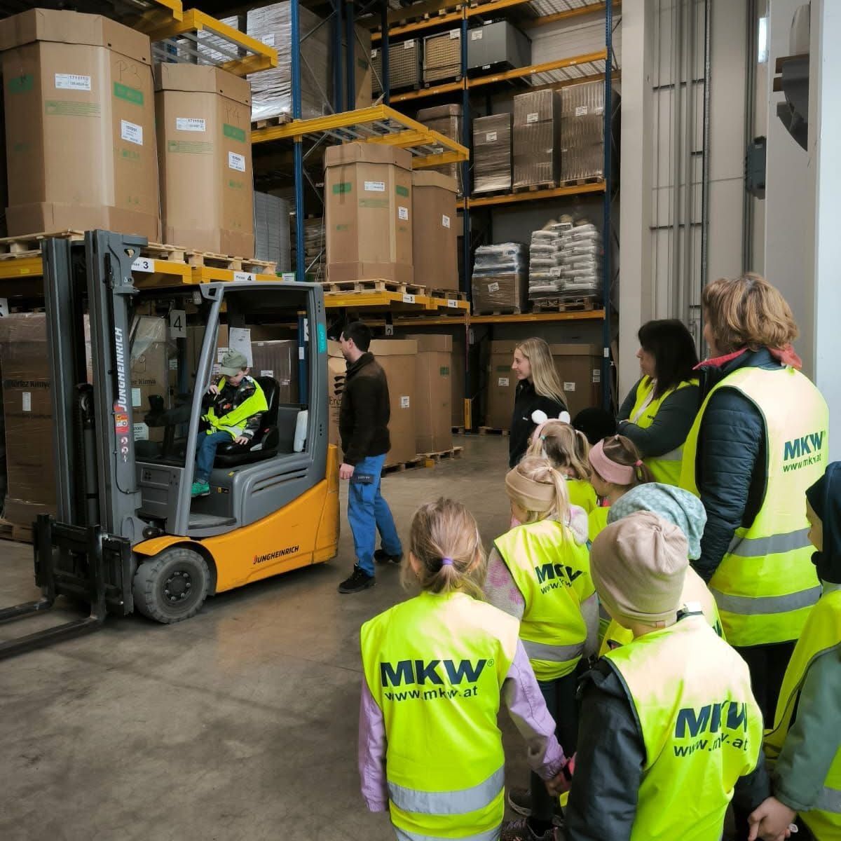 Eine Gruppe von Kindern und Erwachsenen in Sicherheitswesten beobachtet einen Gabelstaplerfahrer in einem Lager, das mit gestapelten Kisten gefüllt ist.