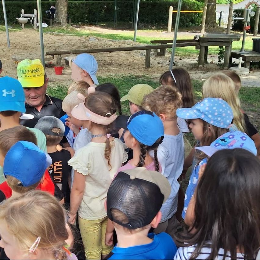 Eine Gruppe von Kindern und Erwachsenen versammelt sich in einem Outdoor-Spielplatz, wobei ein Mann mit einer Mütze mit dem Wort Hermann darauf die Gruppe anführt.