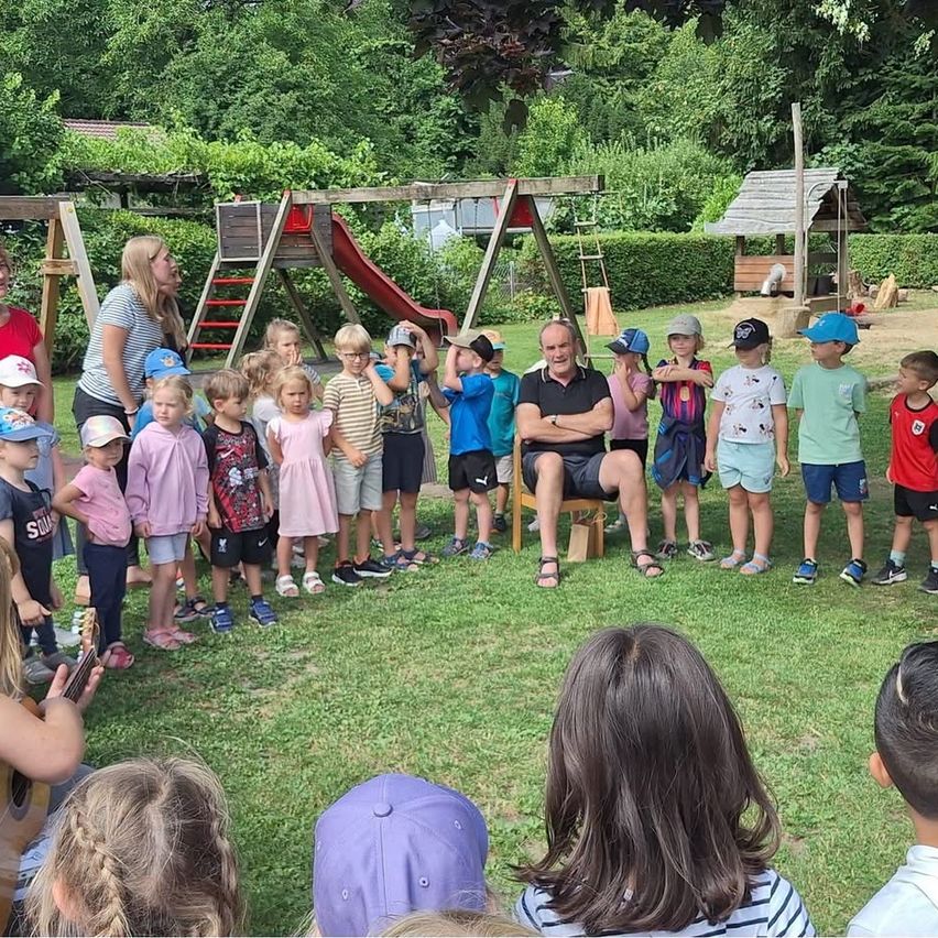 Eine Gruppe von Kindern und Erwachsenen versammelt sich auf einem Rasen, möglicherweise für eine Schulveranstaltung. Ein Mann sitzt auf einem Stuhl, und eine Frau steht in der Nähe der Rutsche.