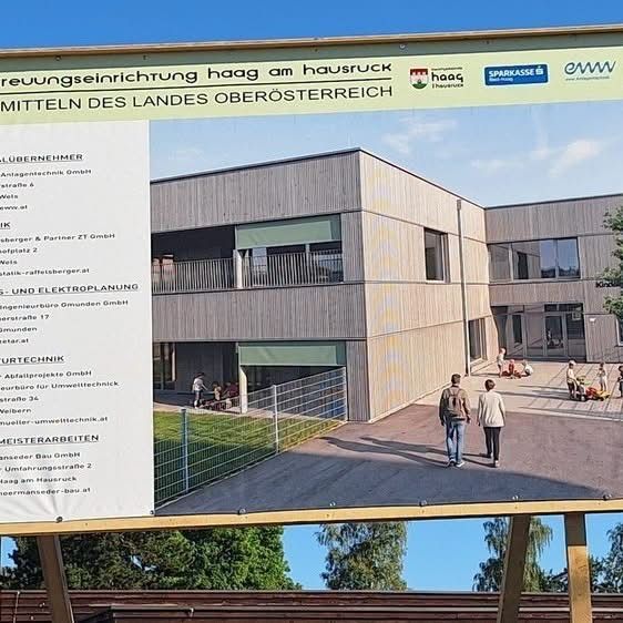 Ein großes Schild zeigt Informationen über ein Bauprojekt in Hausruck, Österreich. Das Schild enthält Logos, Firmennamen und ein Foto des Gebäudes. Menschen gehen um das Gebäude herum.