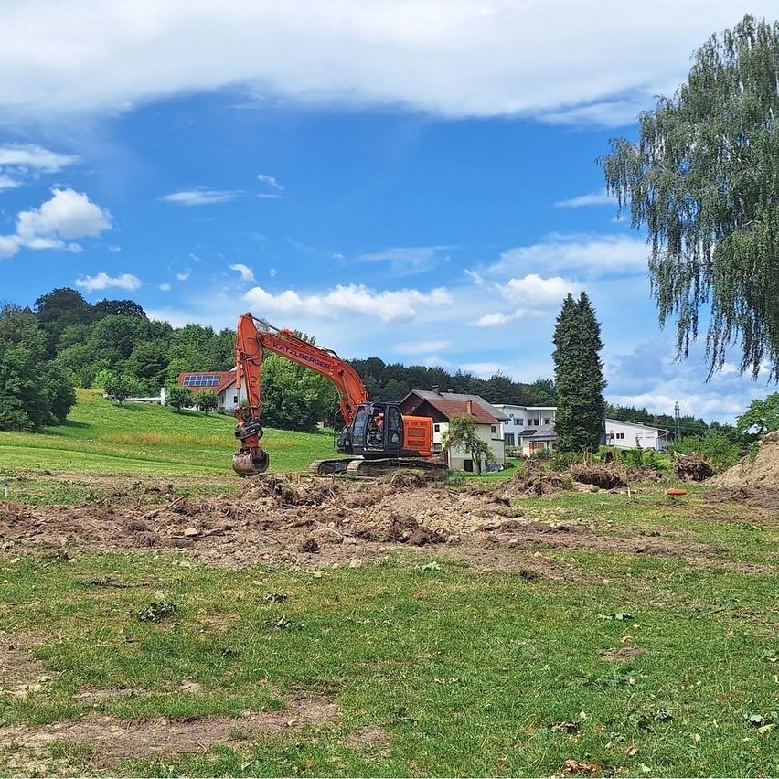 Ein orangefarbener Bagger gräbt den Boden in einem Grasfeld mit Häusern und Bäumen im Hintergrund aus.