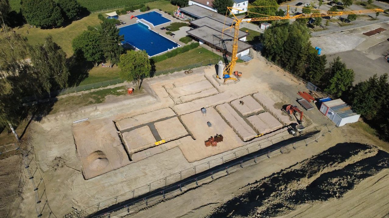 Eine Luftaufnahme einer Baustelle mit einem großen Schwimmbad, einem Kran und verschiedenen Baumaschinen. Die Baustelle ist von Bäumen und Gras umgeben.