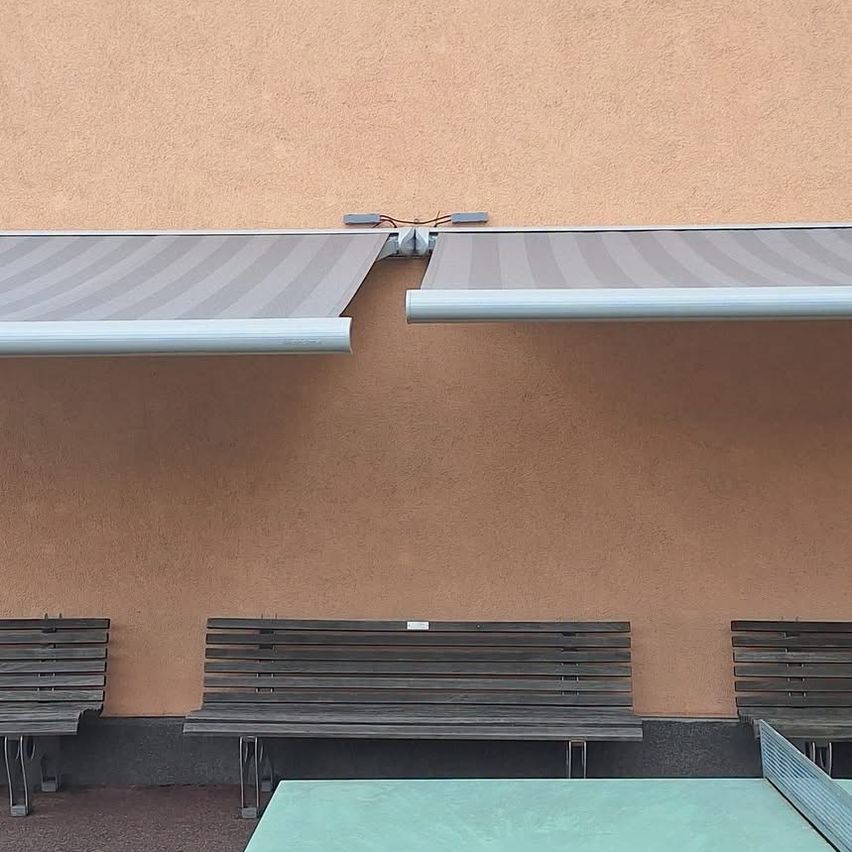 Eine beige Wand mit einem grauen Vordach über drei Holzbänken und einem grünen Tischtennistisch.