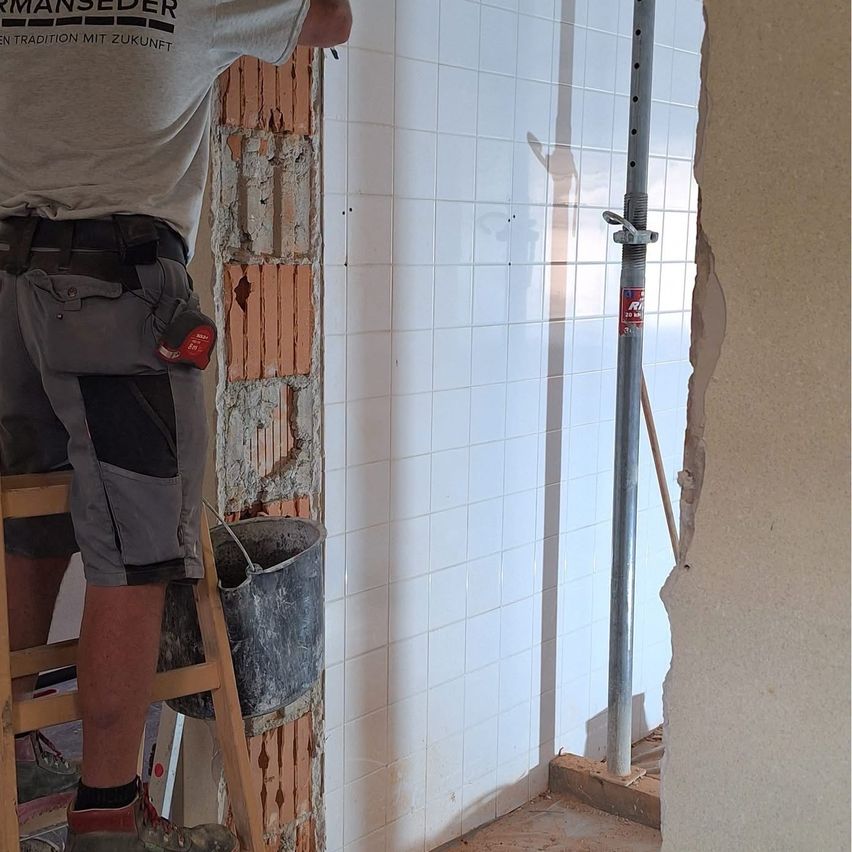 Ein Bauarbeiter steht auf einer Leiter, trägt ein graues Shirt und Shorts und hält ein Werkzeug. Er arbeitet an einer Wand mit freiliegenden Ziegeln und einem Eimer in der Nähe.