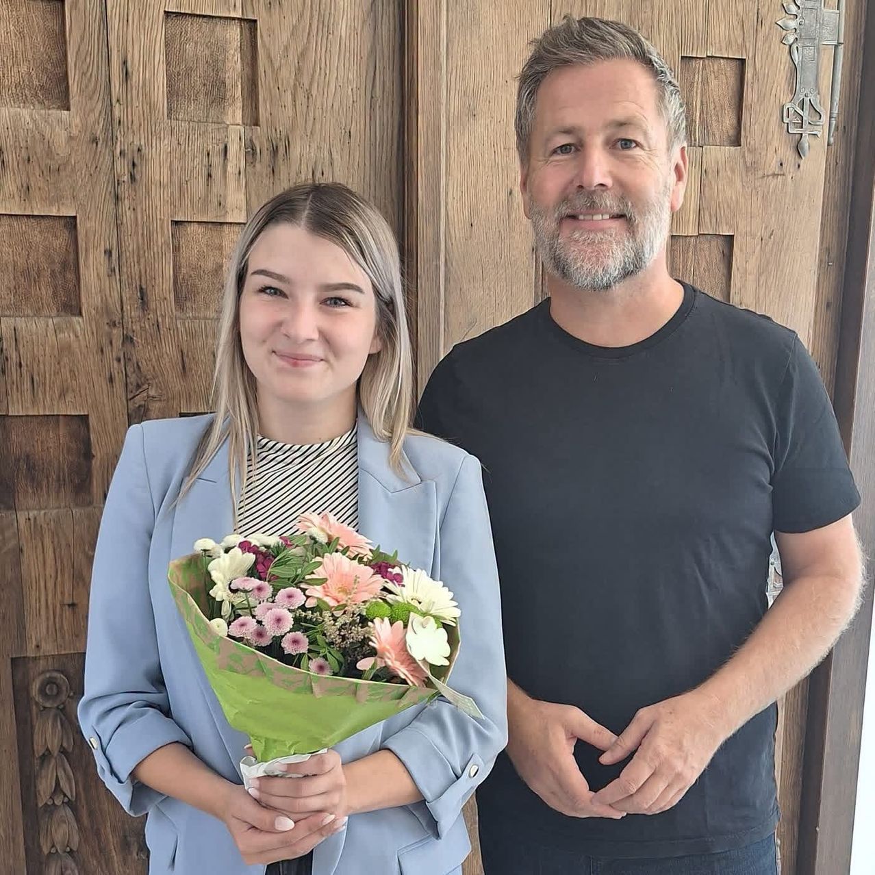 Ein Mann und eine Frau stehen lächelnd vor einer Holztür. Die Frau hält einen Blumenstrauß.
