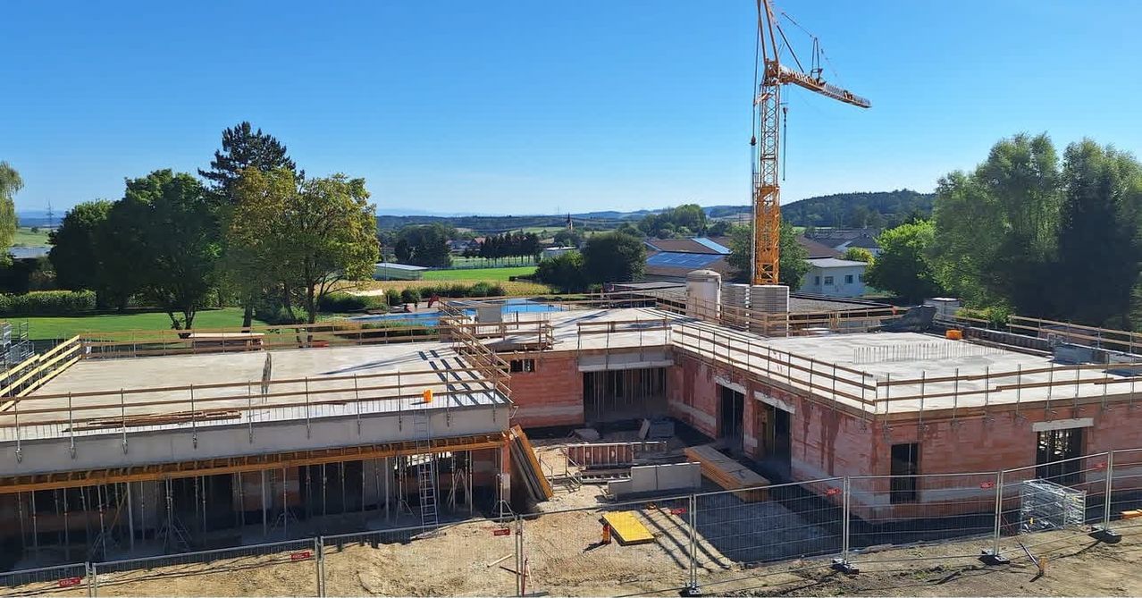 Luftaufnahme einer Baustelle mit einem Kran, umgeben von Bäumen und einem klaren blauen Himmel. Das im Bau befindliche Gebäude hat einen Pool auf dem Dach.
