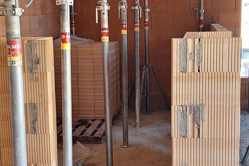 Ein Lager mit Metallstangen und auf dem Boden gestapelten Holzpaletten. Die Stangen haben Etiketten und die Wände bestehen aus Ziegelsteinen.