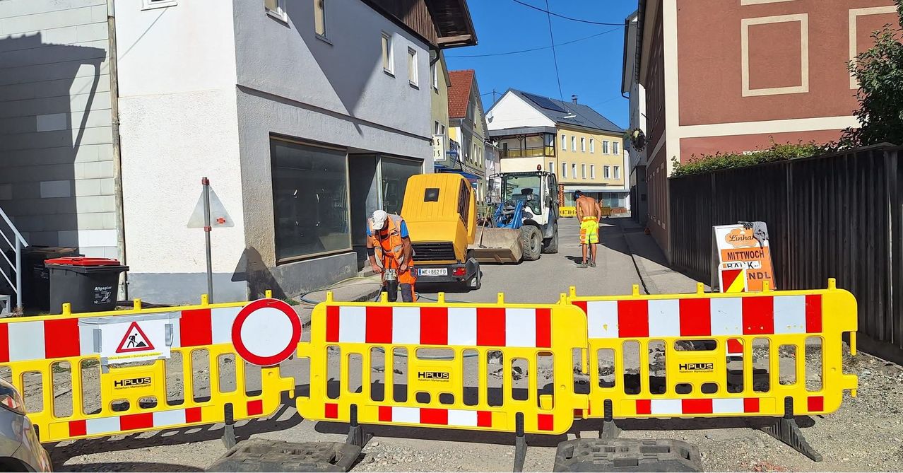 Ein Bauarbeiter reinigt die Straße mit einer Maschine, während ein anderer Arbeiter in der Nähe geht. Die Straße ist mit Absperrungen blockiert.