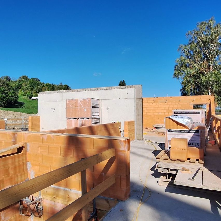 Ein Bauplatz mit orangefarbenen Ziegeln und einem klaren blauen Himmel. Holzstrukturen und einige Kisten sind sichtbar.