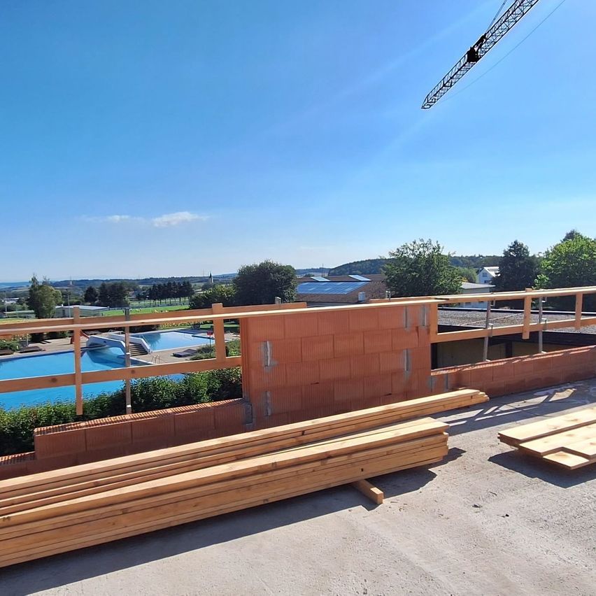 Ein Bauplatz mit Holzbalken und einem Pool im Hintergrund unter einem klaren Himmel.