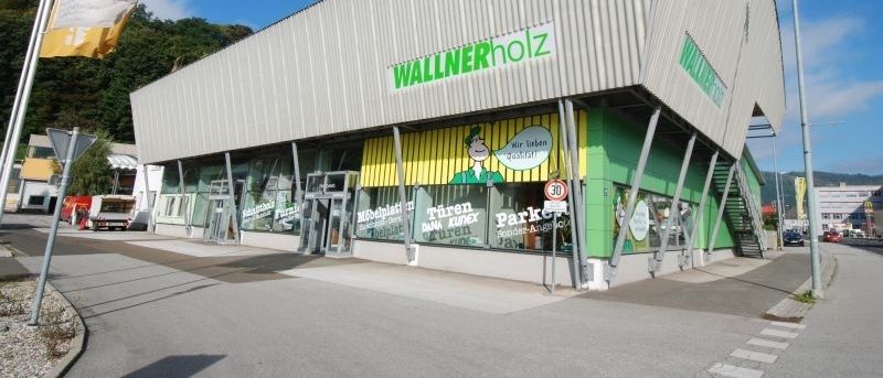 Ein Geschäft mit einer grünen Wand und dem Namen Wallner Holz befindet sich an der Straße. Es sind einige Autos auf der Seite geparkt.