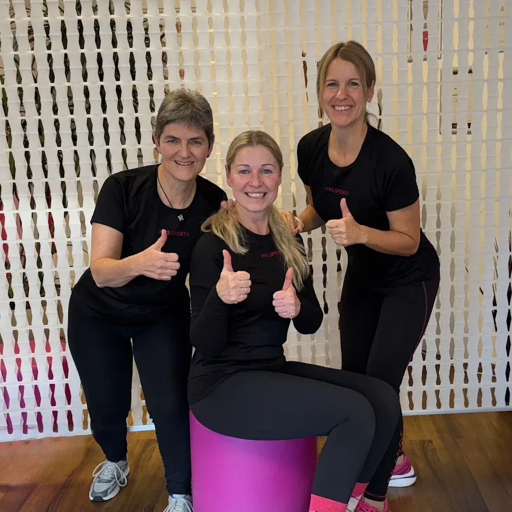 Drei Frauen lächeln und posieren für ein Foto. Die Frau in der Mitte sitzt auf einem lila Hocker und gibt einen Daumen hoch. Die Frau auf der linken Seite steht hinter ihr und gibt auch einen Daumen hoch. Die Frau auf der rechten Seite steht neben der Frau in der Mitte und gibt ebenfalls einen Daumen hoch. Sie tragen alle schwarze T-Shirts und schwarze Hosen.