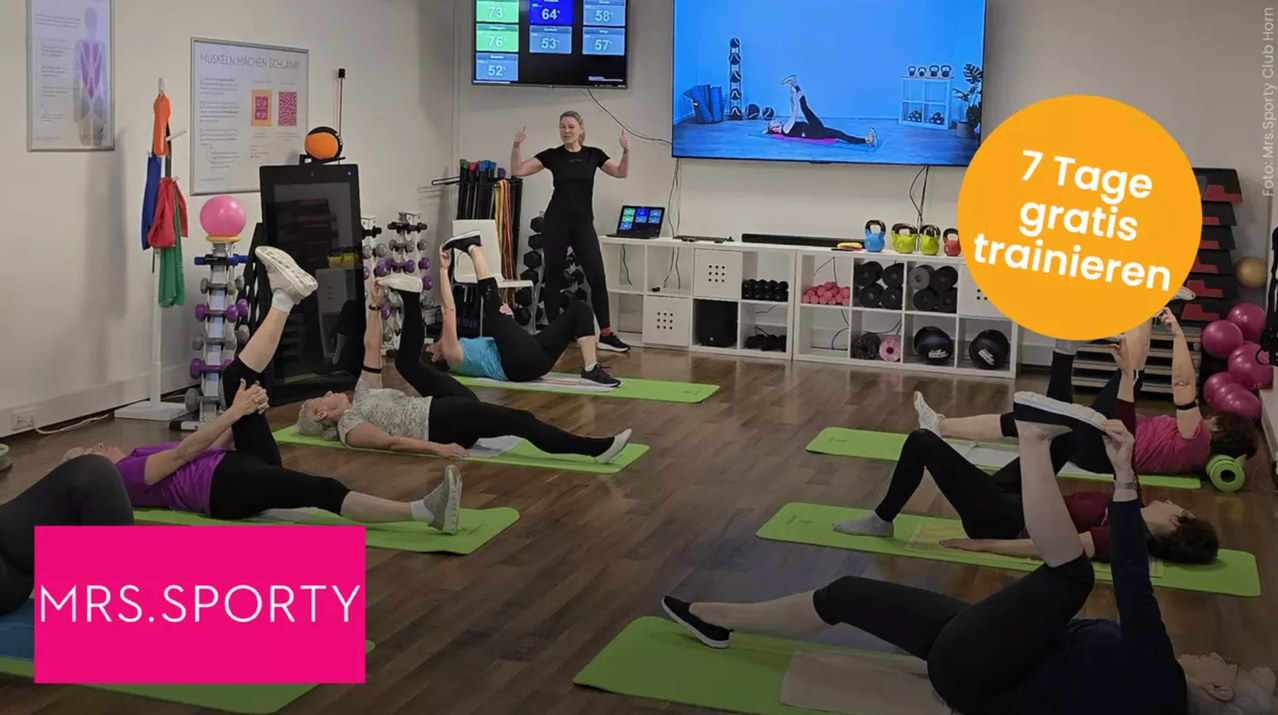 Mehrere Personen machen Yoga auf grünen Yogamatten. Eine Frau leitet den Unterricht, und auf dem Bildschirm an der Wand ist eine andere Frau zu sehen, die Yoga macht. Verschiedenes Trainingsausrüstung ist auf den Regalen aufgestellt.