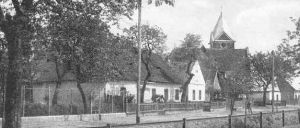 Ein Schwarz-Weiß-Foto zeigt eine Dorfstraße mit traditionellen Häusern, eines davon mit einem Kirchturm, umgeben von Bäumen und einem Zaun.