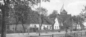 Ein Schwarz-Weiß-Foto zeigt eine Dorfstraße mit traditionellen Häusern, eines davon mit einem Kirchturm, umgeben von Bäumen und einem Zaun.