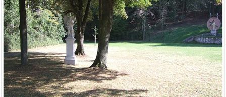 Ein ruhiger Park mit einer Statue unter einem Baum, umgeben von üppigem Grün und einem Rasenfeld.