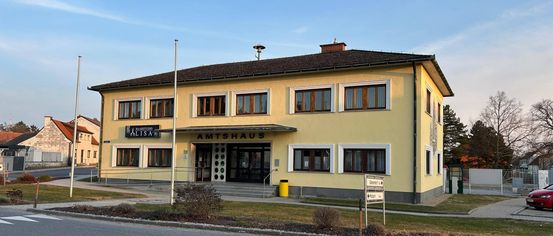 Ein gelbes Gebäude mit mehreren Fenstern und einem braunen Dach, beschriftet mit 'Amtshaus', steht vor einer Straße mit einem Straßenschild.
