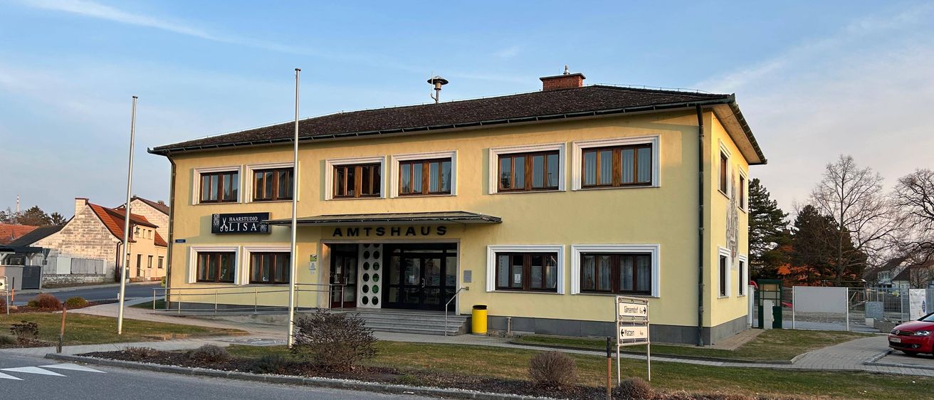 Ein gelbes Gebäude mit mehreren Fenstern und einem braunen Dach, beschriftet mit 'Amtshaus', steht vor einer Straße mit einem Straßenschild.