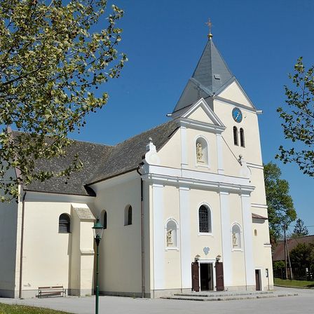 Eine weiße Kirche mit Turm und Uhr steht unter blauem Himmel. Die Fassade hat bogenförmige Fenster und Statuen, davor steht eine Bank.