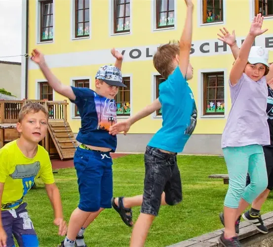 Eine Gruppe von Kindern springt mit erhobenen Händen vor einem gelben Gebäude mit Fenstern. Dahinter befindet sich ein Rasenbereich mit einer Holzspielanlage.