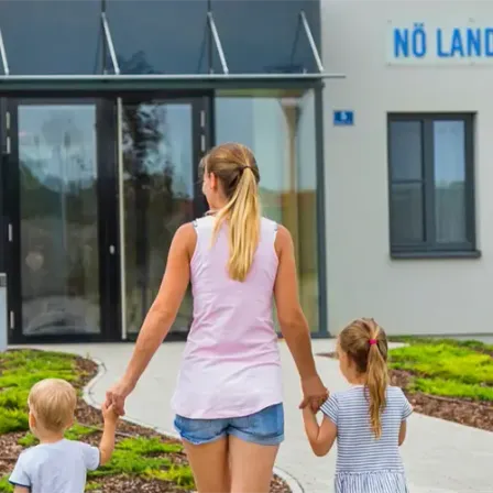 Eine Frau und zwei Kinder gehen vor einem Gebäude mit einem Schild, auf dem 'Keine Landeskinder' steht. Das Gebäude hat eine Glastür und Fenster.