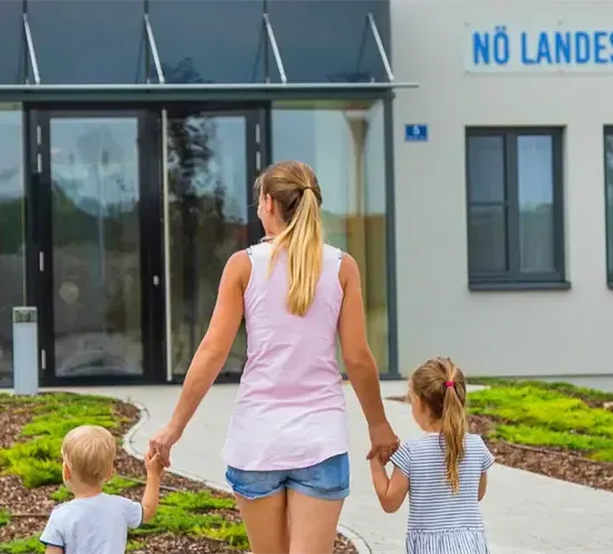 Eine Frau und zwei Kinder gehen vor einem Gebäude mit einem Schild, auf dem 'Keine Landeskinder' steht. Das Gebäude hat eine Glastür und Fenster.