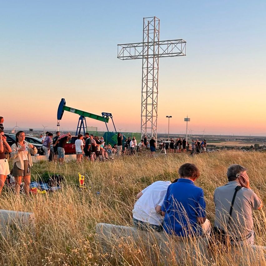 Eine Menschenmenge versammelt sich um ein großes Kreuz auf einem Feld, mit einem Sonnenuntergang im Hintergrund und einer Pumpe in der Nähe.