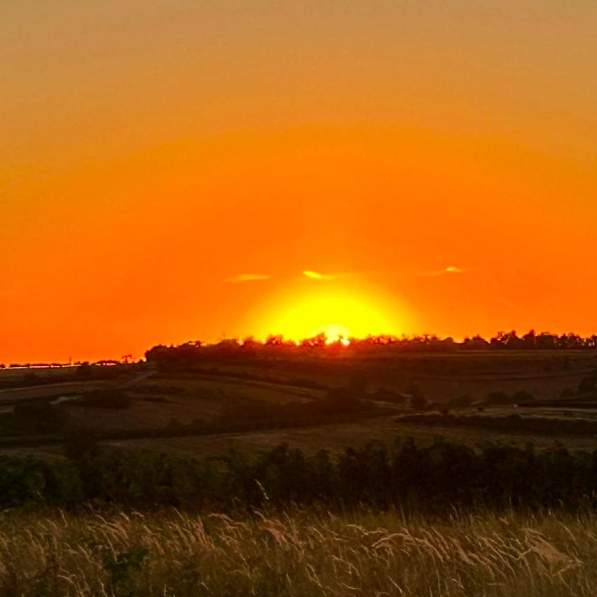 Sonnenuntergang über einem weiten, offenen Feld mit einer Horizontlinie, Bäumen und einer kleinen Silhouette eines Gebäudes.