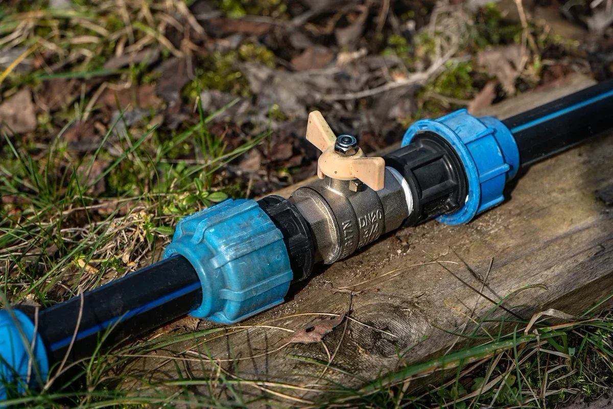Ein blauer und messingfarbener Gartenschlauchverbinder liegt auf einem Holzstamm im Freien, mit grünem Gras und Moos im Hintergrund.