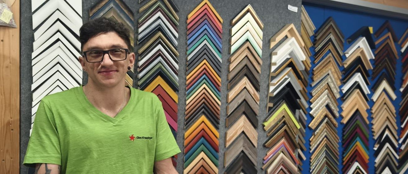 Ein Mann in einem grünen T-Shirt steht vor einer Wand mit farbenfrohen Bilderrahmen. Er lächelt und hat seine Hände auf einem Tisch mit puderzuckerbedeckten Donuts.