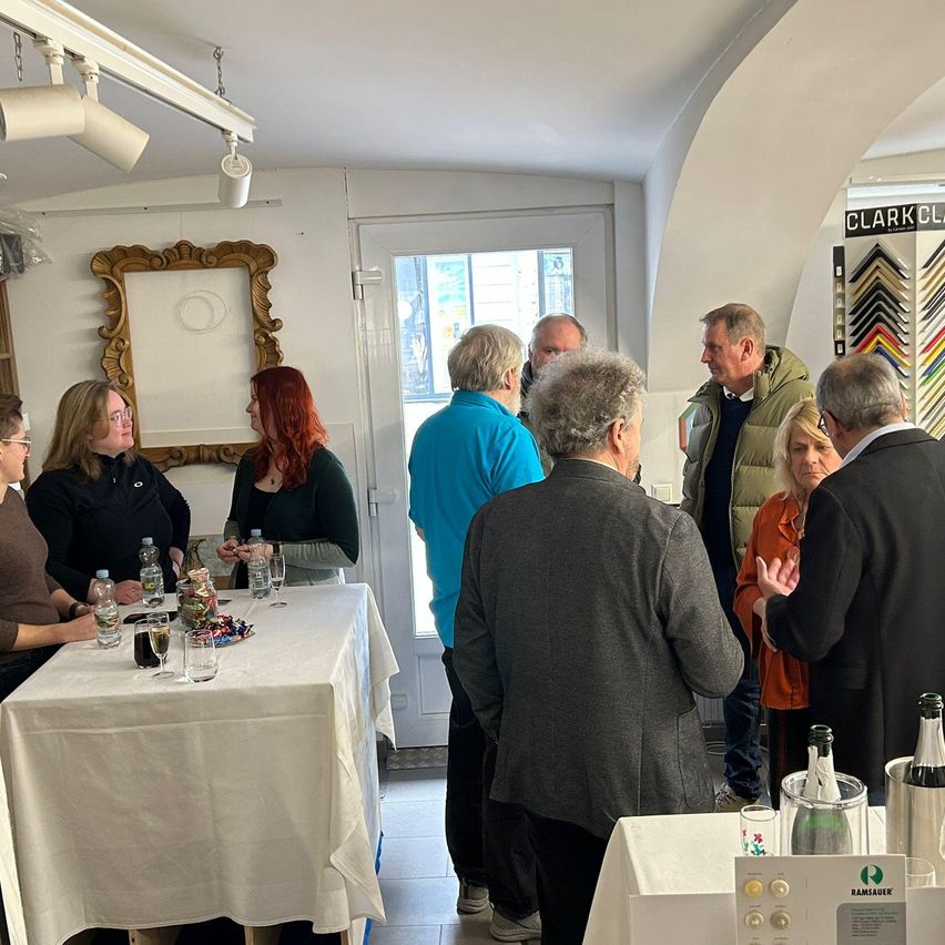 Eine Gruppe von Menschen steht um Tische in einem Raum mit weißen Wänden. Im Hintergrund befindet sich eine Tür. Flaschen, Gläser und Süßigkeiten sind auf den Tischen.