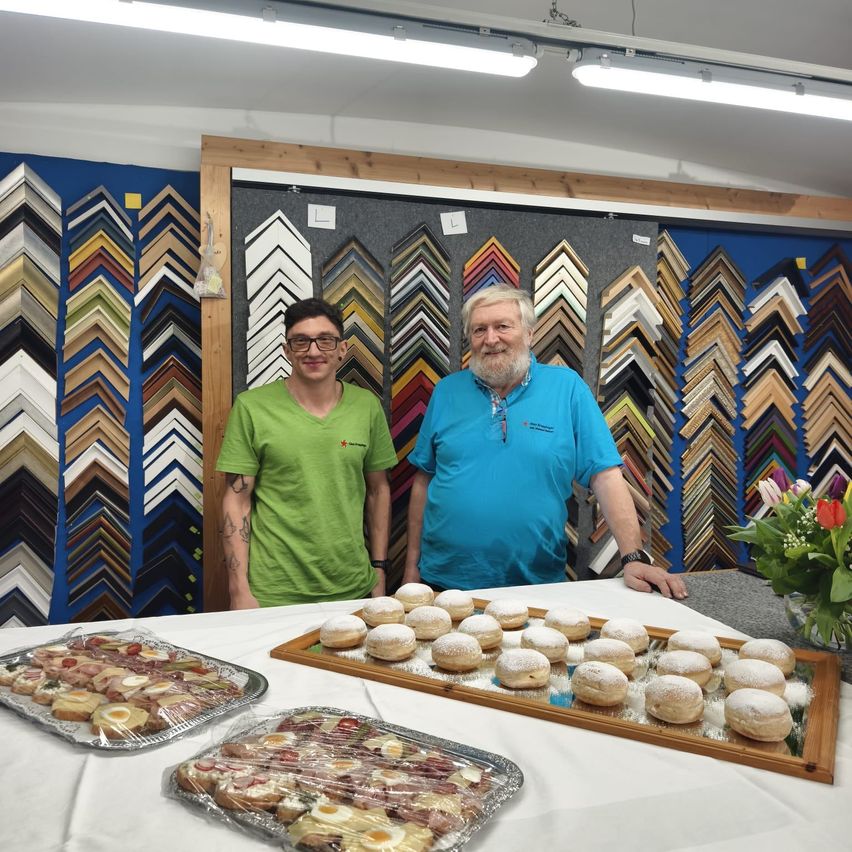 Zwei Männer stehen hinter einem Tisch voller Essen. Einer trägt ein grünes T-Shirt, der andere ein blaues. Sie stehen vor einer Wand mit verschiedenen Bilderrahmen.