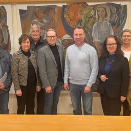 Sieben Personen stehen gemeinsam vor einer großen Wandmalerei. Das Wandbild zeigt verschiedene Figuren in leuchtenden Farben. Jede Person trägt formelle Kleidung, was auf eine offizielle Veranstaltung hindeutet.