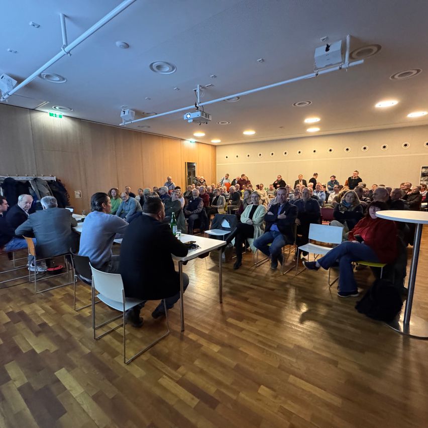 Ein Raum mit Holzboden beherbergt eine Versammlung von Menschen. Einige sitzen an Tischen, andere in Stühlen. Ein Projektor und Deckenleuchten sind sichtbar.